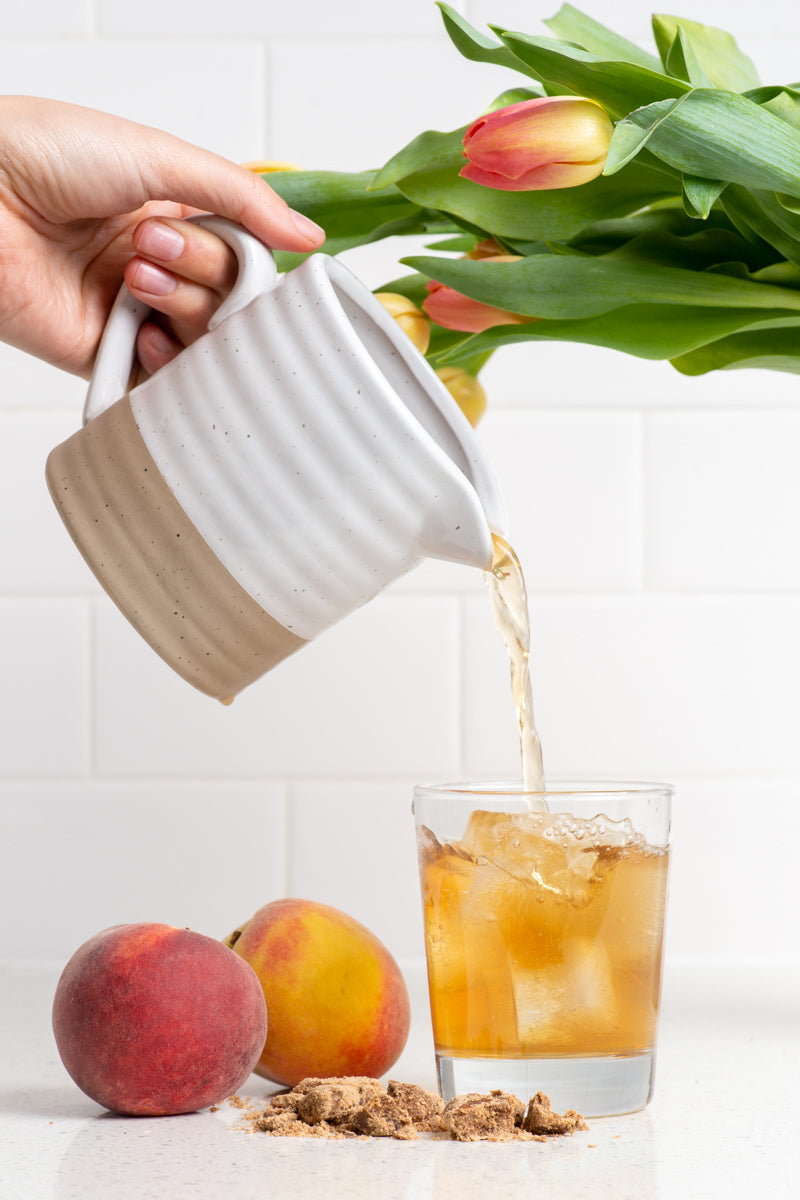 A hand pouring tea from a ceramic pitcher into a glass with ice, on a counter with 2 peaches, a pile of brown sugar, and tulip flowers. The tea is Iced Sweet Peach from True Tea, a tea company based in Portland, Oregon