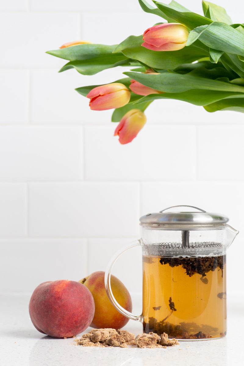 Glass tea brewer on a counter with a pile of brown sugar, 2 peaches, and tulip flowers. The tea is Iced Sweet Peach from True Tea, a tea company based in Portland, Oregon